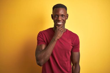 Kırmızı t-shirt giyen genç Afro-Amerikan bir adam izole edilmiş sarı arka planda dikiliyor kameraya bakıyor kollarını kavuşturup, ellerini çenesine kaldırarak gülümsüyor. Olumlu düşünüyorum..