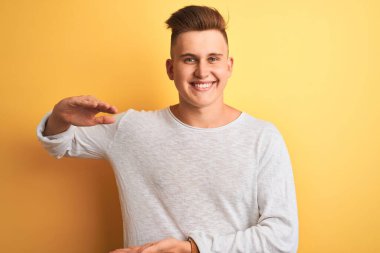 İzole edilmiş sarı arka plan üzerinde duran beyaz tişörtlü genç yakışıklı adam. Büyük ve büyük boyut işareti, ölçü sembolü gösteren el hareketleriyle. Kameraya bakıp gülümsüyor. Ölçüm konsepti.