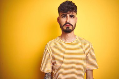 İzole edilmiş sarı arka plan üzerinde duran çizgili tişörtlü genç adam depresif ve endişeli, sinirli ve korkmuş. Üzücü ifade.