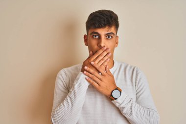 Young indian man wearing sweater standing over isolated white background shocked covering mouth with hands for mistake. Secret concept.