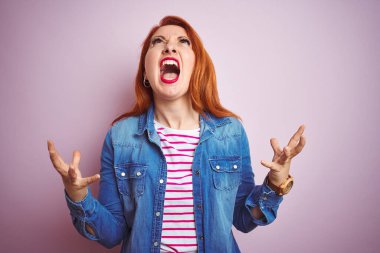 Güzel kızıl saçlı kadın kot gömlek ve çizgili tişört giyiyor pembe arka planda çılgınca bağırıyor ve agresif bir ifade ve kollarla bağırıyor. Hayal kırıklığı konsepti.