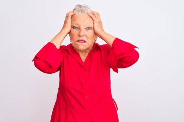 Yaşlı, gri saçlı, kırmızı gömlekli bir kadın izole edilmiş beyaz arka planda dikiliyor. Baş ağrısı, acı ve migren yüzünden stres altında. Ellerini başının üstüne koy..