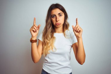 İzole edilmiş arka plan üzerinde günlük beyaz tişört giyen genç ve güzel bir kadın üzgün ve üzgün görünüyor, parmaklarıyla yönü gösteriyor, mutsuz ve depresif..