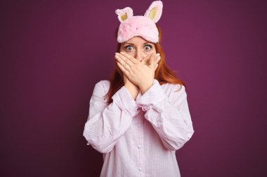 Genç, güzel, kızıl saçlı, pijama giyen ve mor izole edilmiş arka planda maske takan bir kadın ağzını yanlışlıkla ellerle kapatarak şok etti. Gizli konsept.