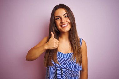 İzole edilmiş pembe arka planda duran çizgili elbise giyen genç ve güzel bir kadın mutlu bir el hareketi yapıyor. Kameraya bakıp başarıyı gösteren ifade onaylanıyor.