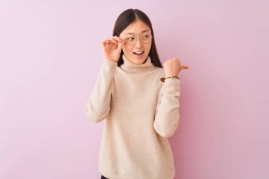 Balıkçı yaka kazak ve gözlük takan genç Çinli bir kadın izole edilmiş pembe arka plan üzerinde mutlu bir gülümsemeyle baş parmağıyla işaret ediyor.