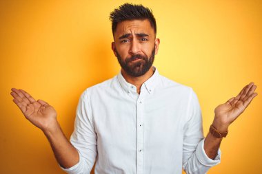 İzole edilmiş sarı arka plan üzerinde duran beyaz şık gömlekli Hintli işadamı elleri ve kolları havada, kafası karışık bir ifade takınıyor. Şüphe kavramı.
