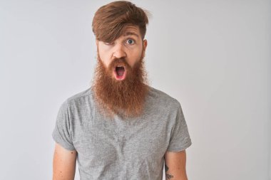 Genç kızıl İrlandalı adam izole gri arka plan üzerinde duran t-shirt giyen korkmuş ve sürpriz ifade, korku ve heyecanlı yüz ile şok.