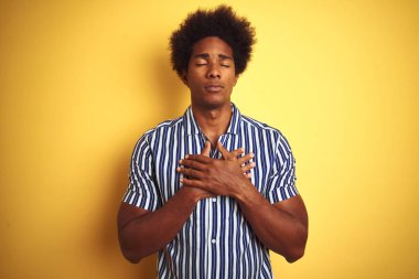 Afro saçlı, çizgili tişörtlü, izole edilmiş sarı arka plan üzerinde duran, elleri göğsünde, gözleri kapalı ve yüzünde minnet dolu bir jest olan Amerikalı bir adam. Sağlık kavramı.