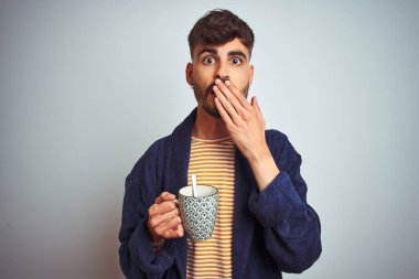 Bornoz giyen genç adam izole beyaz arka plan kapağı ağız üzerinde duran kahve içme el hata için utanç ile şok, korku ifadesi, sessizlik, gizli kavram korkmuş