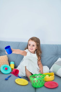 Sevimli sarışın bebek kanepede oturuyor evde yemek ve çatal bıçakla yemek pişiriyor.