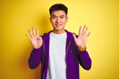 Mor kazak giyen Asyalı genç adam izole edilmiş sarı arka planda duruyor ve dokuz numarayı işaret ediyor. Bu arada kendinden emin ve mutlu gülümsüyor..