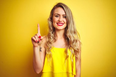 Sarı arka planda duran tişört giyen genç saldırgan kadın bir numaralı parmağı gösterip işaret ederken kendinden emin ve mutlu bir şekilde gülümsüyor..