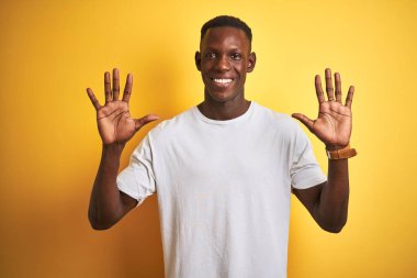 Beyaz tişörtlü genç Afro-Amerikalı adam izole edilmiş sarı arka plan üzerinde duruyor ve parmaklarını on numarayı gösterirken kendinden emin ve mutlu bir şekilde gülümsüyor..