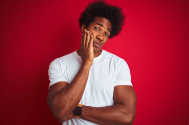 Siyah saçlı, beyaz tişörtlü, izole edilmiş kırmızı arka planda duran genç Amerikalı adam, çapraz kollu depresyondan sıkılmış ve yorgun göründüğünü düşünüyor..
