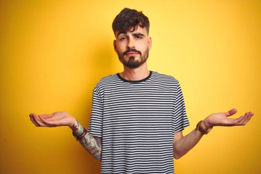 Çizgili t-shirt giyen dövme giyen genç adam izole sarı arka plan üzerinde duran habersiz ve kolları ve elleri ile karışık ifade kaldırdı. Şüphe kavramı.