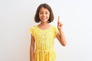 Sarı çiçekli elbise giyen güzel bir kız çocuğu izole edilmiş beyaz arka plan üzerinde duruyor ve bir numaralı parmağını gösterip gülümserken kendinden emin ve mutlu.