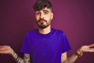 İzole edilmiş mor arka plan üzerinde duran dövmeli genç bir adam. Kolları ve elleri havada, kafası karışmış bir ifade. Şüphe kavramı.