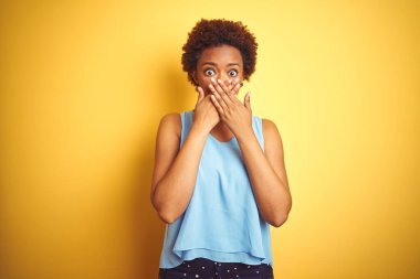 Zarif bir tişört giyen güzel Afro-Amerikalı kadın izole edilmiş sarı arka planda ağzı yanlışlıkla ellerle kapatarak şok geçirdi. Gizli konsept.