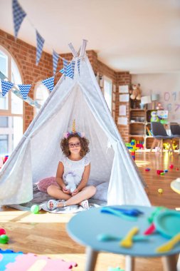 Gözlüklü ve tek boynuzlu at diademi takmış güzel bir çocuk Tip 'in içinde yerde oturuyor ve anaokuluna gülümsüyor.