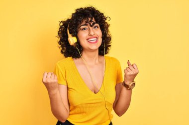 Kıvırcık saçlı, kulaklık takan, izole edilmiş sarı arka planda müzik dinleyen Arap bir kadın kollarını kaldırarak ve açık gözlerle başarıyı kutluyor. Kazanan konsept.