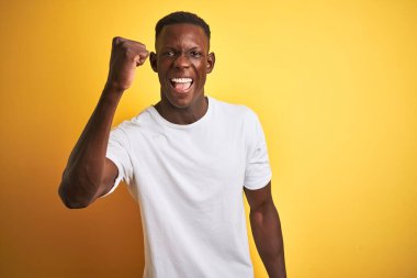 Beyaz t-shirt giyen genç Afro-Amerikan bir adam izole edilmiş sarı arka planda kızgın ve kızgın duruyor öfkeyle bağırırken yumruğunu kaldırıyor. Öfke ve agresif kavram.