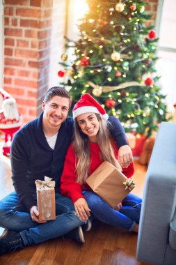 Mutlu ve kendinden emin gülümseyen genç bir çift. Elinde hediyelerle yerde oturup Noel ağacının etrafında sarılmak.