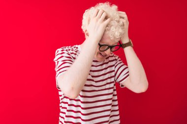 Sarı çizgili tişört ve gözlük takan genç albino adam izole edilmiş kırmızı arka plan üzerine çaresiz ve stresli baş ağrısı ve migren yüzünden acı çekiyor. Ellerini başının üstüne koy..