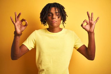 Rastgele tişört giyen Afro bir adam izole edilmiş sarı arka planda dikiliyor rahatlıyor ve gözleri kapalı meditasyon yaparken parmaklarıyla gülümsüyor. Yoga konsepti.