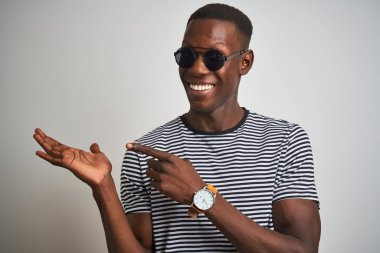 İzole edilmiş beyaz arka plan üzerinde çizgili tişört ve güneş gözlüğü takan Afrikalı Amerikalı adam kameraya hayret ediyor ve gülümsüyor..
