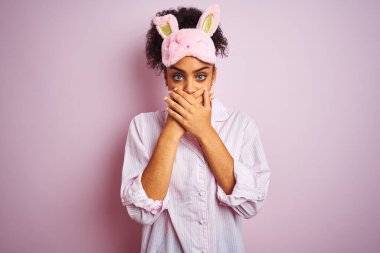 Genç Afro-Amerikalı kadın pijama giyiyor ve pembe arka planda maske takıyor. Ağzını yanlışlıkla ellerle kapatıyor. Gizli konsept.