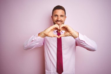 İş adamı, pembe izole edilmiş arka plan üzerinde kravat ve şık gömlek takıyor. Aşkla gülümsüyor. Elleriyle kalp sembolü ve şekli gösteriyor. Romantik konsept.