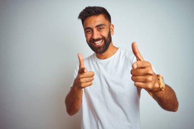 İzole edilmiş beyaz arka planda duran tişört giyen genç Hintli adam mutlu ve komik yüzlü parmaklarını kameraya doğrultuyor. İyi enerji ve titreşimler.