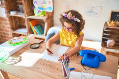 Gözlüklü ve tek boynuzlu at diademi takmış güzel bir çocuk. Anaokulunda kağıt ve keçeli kalemle çizim yapıyor.