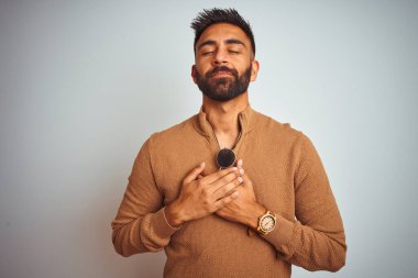 Zarif süveter ve güneş gözlüğü takan Hintli bir adam izole edilmiş beyaz arka planda duruyor elleri göğsünde, gözleri kapalı ve yüzünde minnet dolu bir jest ile gülümsüyor. Sağlık kavramı.