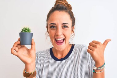 Genç kızıl saçlı kadın, elinde kaktüs saksısını izole arka planda tutuyor ve gülümseyen bir gülümsemeyle baş parmağıyla işaret ediyor.