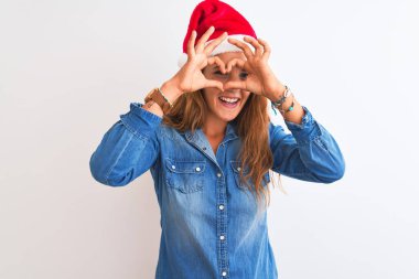 Genç, güzel, kızıl saçlı bir kadın, izole edilmiş bir arka planda Noel şapkası takıyor. El ve parmaklarıyla kalp şeklinde, tabelanın içinden bakıp gülümsüyor.