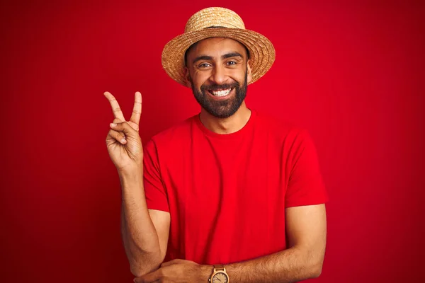 若いハンサムなインド人男性は Tシャツと帽子を着用し カメラで勝利のサインをする幸せな顔をして笑って孤立した赤い背景の上に 第二番 — ストック写真