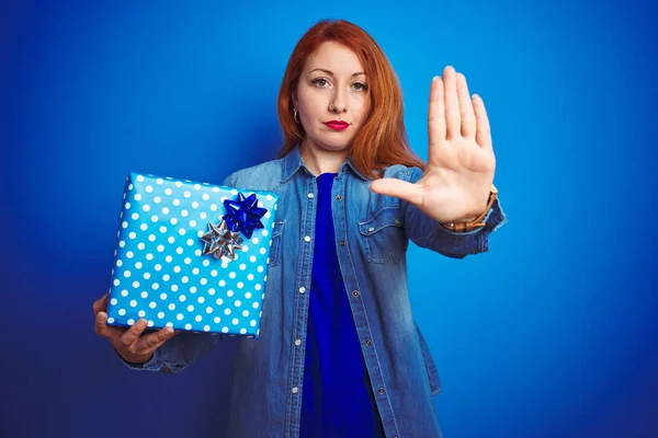Young beautiful redhead woman wearing birthday gift over isolated blue background with open hand doing stop sign with serious and confident expression, defense gesture