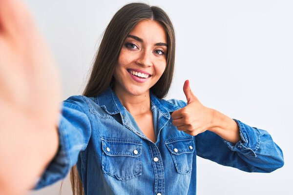 Beautiful woman wearing denim shirt make selfie by camera over isolated white background doing happy thumbs up gesture with hand. Approving expression looking at the camera showing success.