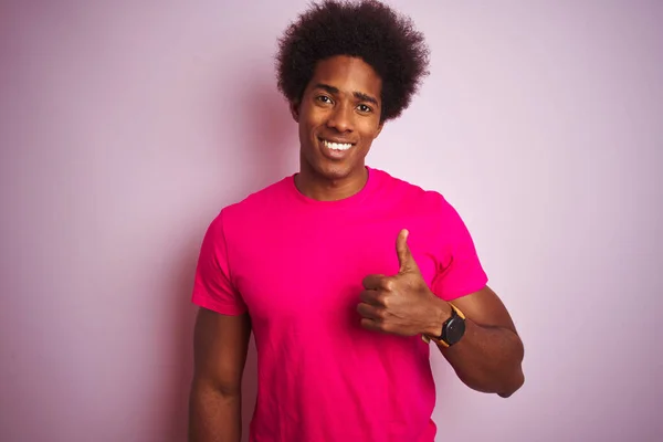 孤立したピンクの背景の上に立ってTシャツを着てアフロ髪を持つ若いアメリカ人男性は 手で幸せな親指アップジェスチャーを行います 成功を示すカメラを見て表現を承認する — ストック写真