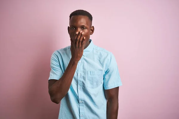 青のカジュアルなシャツを着たアフリカ系アメリカ人男性がピンクの背景に立って手で覆われた口の中で疲れてあくびを退屈 眠れないのか — ストック写真