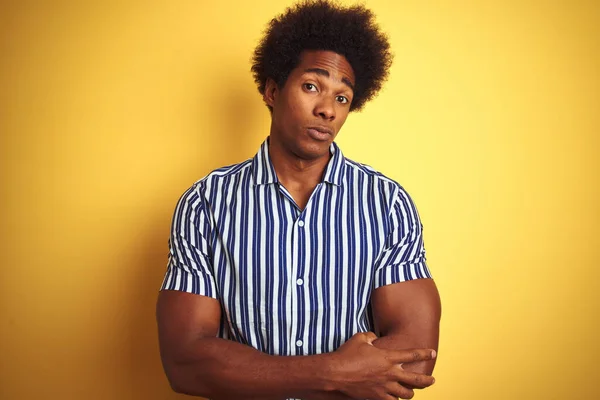 American man with afro hair wearing striped shirt standing over isolated yellow background skeptic and nervous, disapproving expression on face with crossed arms. Negative person.