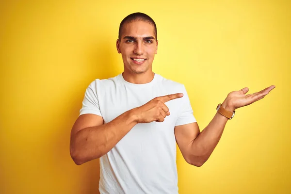 黄色の隔離された背景にカジュアルな白いTシャツを着ている若い白人男性は 手で提示し 指で指している間 カメラに驚いて笑顔 — ストック写真