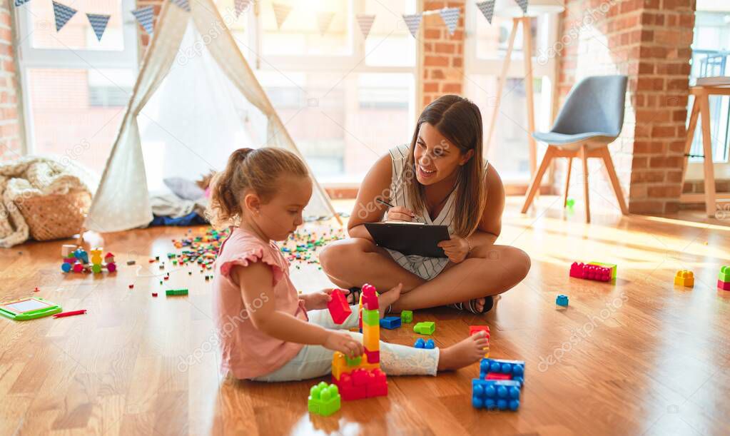 Beautiful psycologist and blond toddler girl doing therapy building tower using plastic blocks at kindergarten