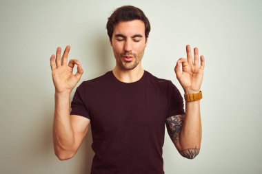İzole edilmiş beyaz arka plan üzerine mor tişört giyen yakışıklı genç adam rahatla ve gözleri kapalı meditasyon hareketi yaparken parmaklarınla gülümse. Yoga konsepti.