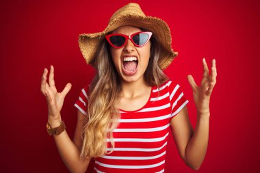 Kırmızı arka plan üzerinde güneş gözlüğü ve yaz şapkası takan genç ve güzel bir kadın sinirli bir şekilde bağırıyor ve bağırıyor. Hayal kırıklığı konsepti.