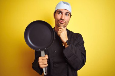 Üniformalı ve şapkalı genç aşçı, izole edilmiş sarı arka planda tava tutuyordu.