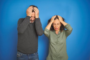 Güzel bir orta yaşlı çift, baş ağrısı, acı ve migren yüzünden çaresiz ve stresli bir şekilde izole edilmiş mavi arka planda duruyorlar. Ellerini başının üstüne koy..