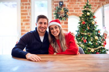 Noel Baba şapkası takan genç bir çift evdeki Noel ağacının etrafındaki sandalyeye ve masaya oturmuş mutlu ve havalı bir gülümsemeyle. Şanslı kişi..